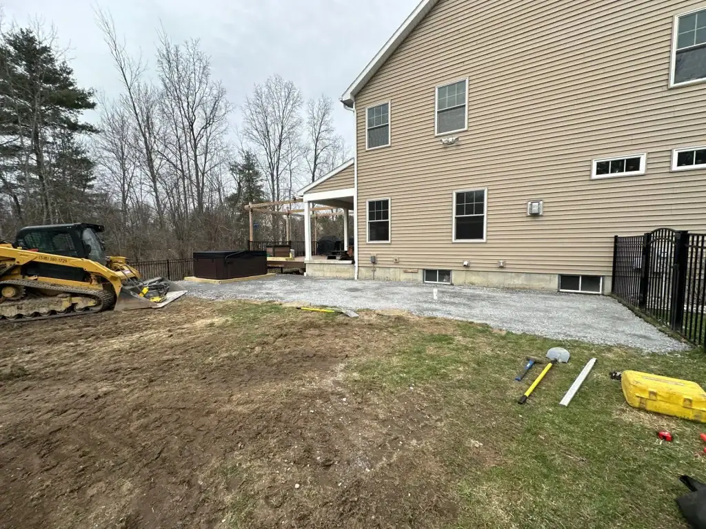 A partially finished backyard patio project beside a tan house in NY, with gravel laid out, tools and equipment from excavating contractors Saratoga County scattered around, a small yellow bulldozer, and a wooden deck in the background amid trees and grass.