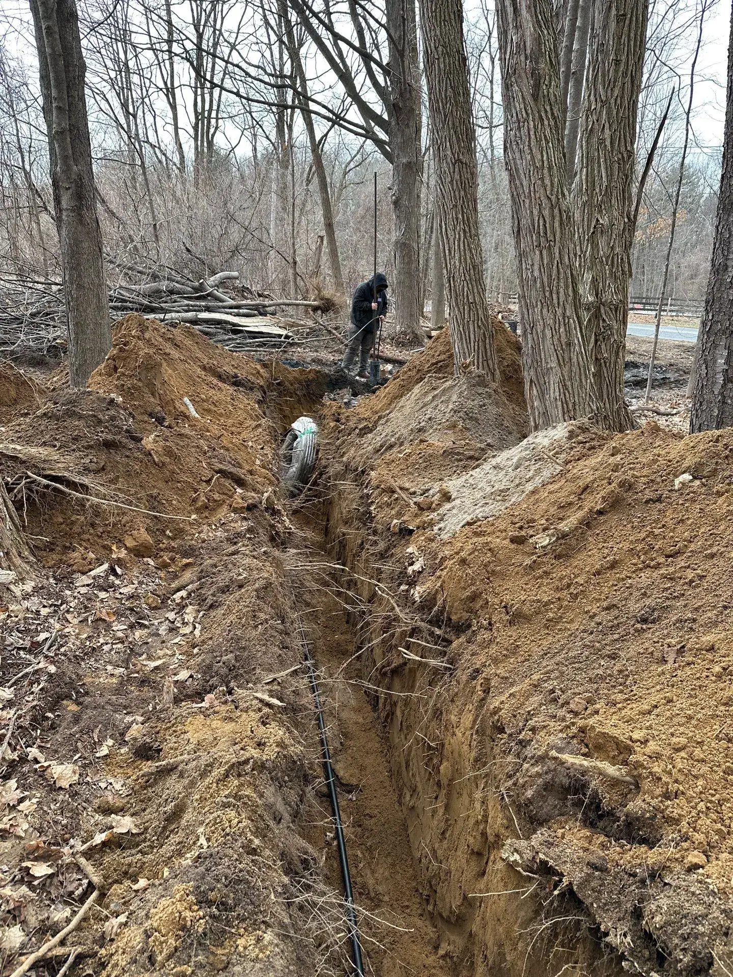 A person stands near a deep trench in a wooded NY area, overseeing another individual working inside the trench where excavating contractors Saratoga County are installing pipes underground among large trees and dug-up earth.