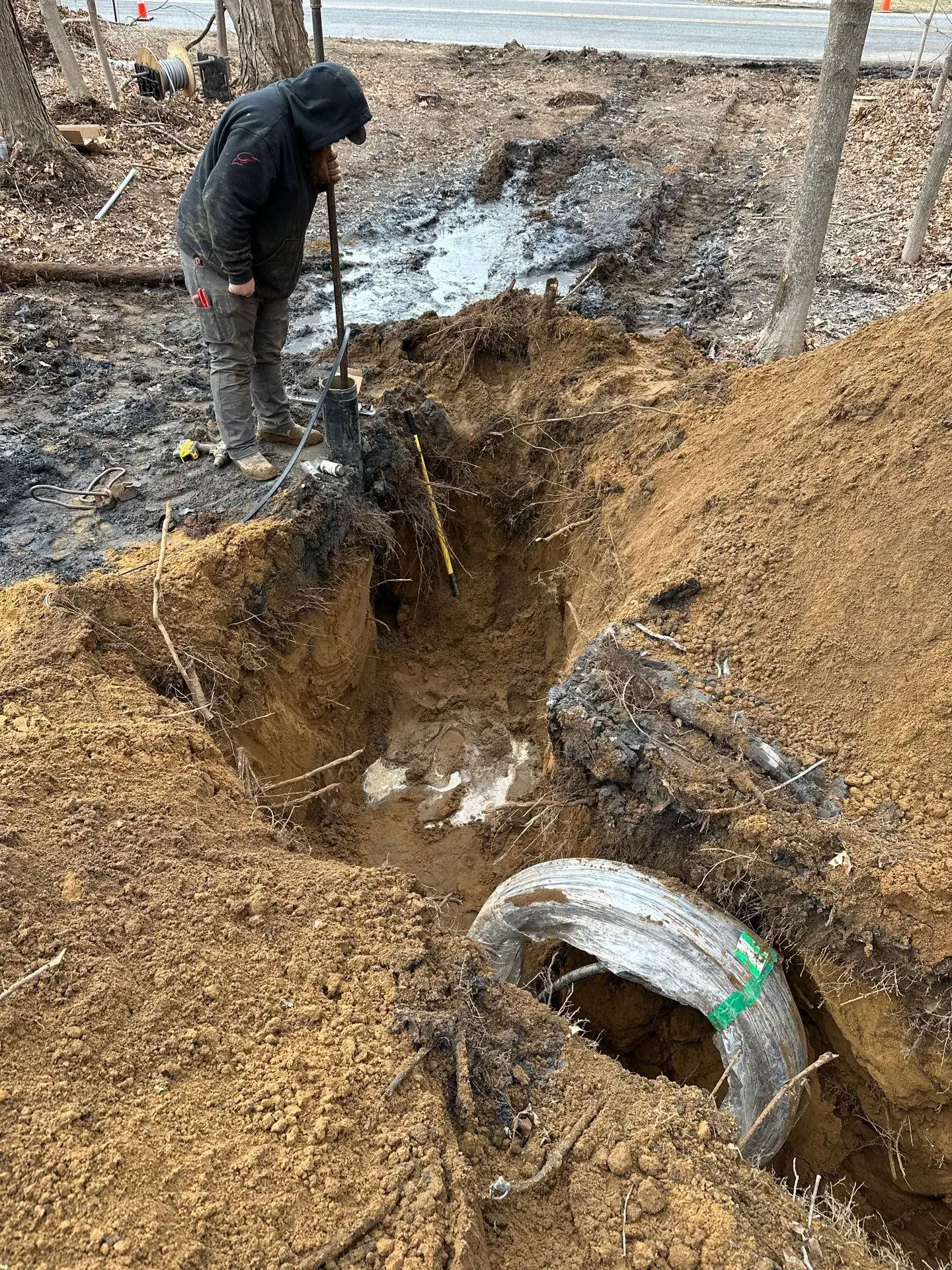 A person wearing a hoodie and boots stands next to a deep, muddy excavation site containing a large, partially buried pipe—evidence of work often handled by excavating contractors in Saratoga County, NY—amid soil and tree roots near a roadside.