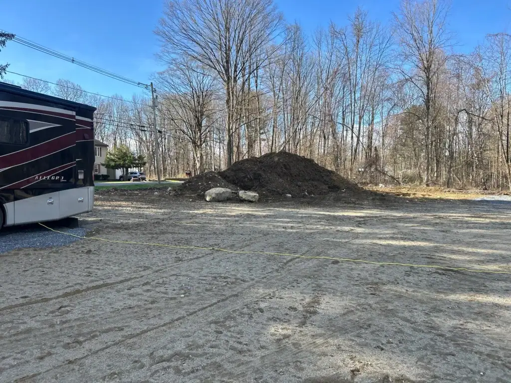 A large pile of dirt and rocks, likely from excavating contractors Saratoga County, NY, sits on bare ground near a wooded area with leafless trees. Part of a maroon and black RV and a house are visible on the left side of the image.
