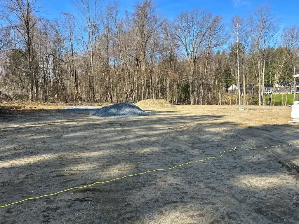 A cleared, dirt lot with piles of gravel and a yellow cord on the ground, likely prepared by excavating contractors Saratoga County, NY. Leafless trees surround the area under a blue sky, with houses partially visible in the background.