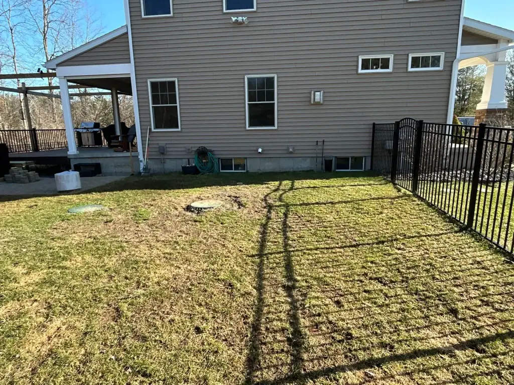 A backyard in NY with patchy grass, a black metal fence casting a shadow, a tan house with several windows, and a covered patio area on the left—perfect for ideas from excavating contractors Saratoga County. A garden hose and utility covers rest against the wall.