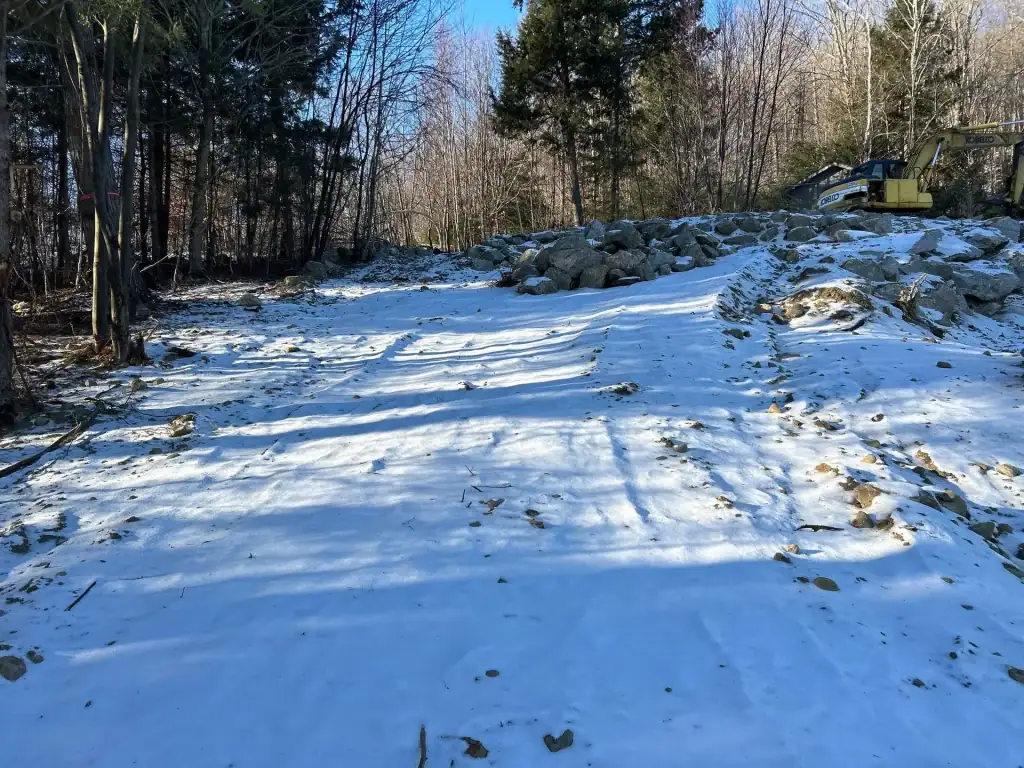A snow-covered clearing in a forest, bordered by trees and large rocks. An excavator from excavating contractors Saratoga County, NY sits at the edge near the rocks under a clear blue sky. Shadows from trees stretch across the snow.