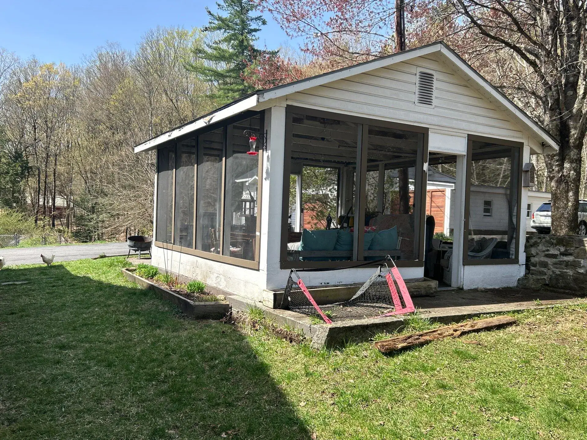 A screened-in porch with patio furniture sits on a grassy NY yard near a road, surrounded by trees and other houses on a sunny day. Pink lawn chairs, a grill, and signs of recent work by excavating contractors Saratoga County are visible outside.