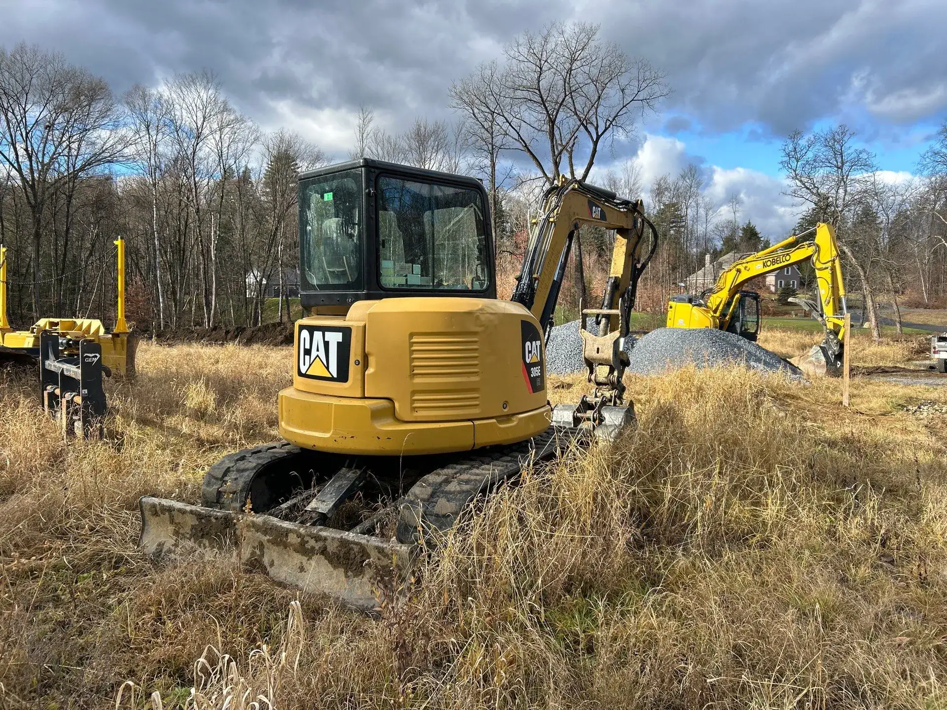 A yellow Caterpillar excavator sits idle in a grassy, open field alongside other construction equipment, showcasing the capabilities of excavating contractors Saratoga County, NY, with bare trees and a cloudy sky in the background.