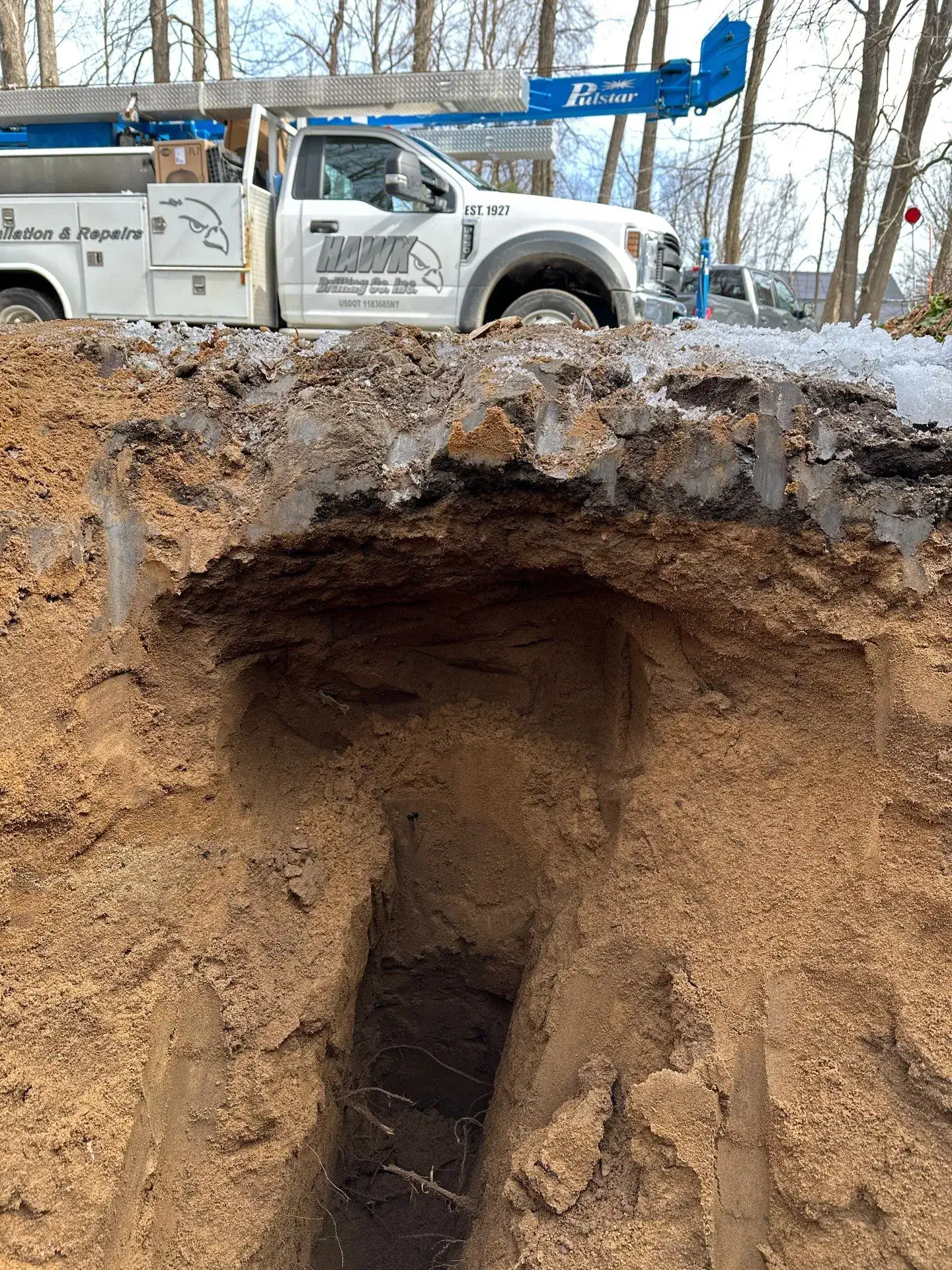 A deep trench dug in sandy soil with roots visible inside, created by excavating contractors Saratoga County, NY; a white utility truck with a crane is parked above on a road lined with leafless trees.