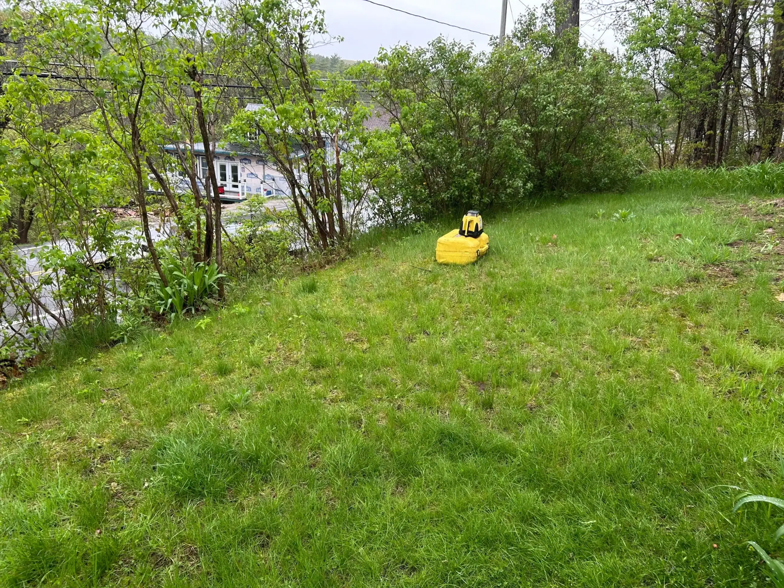 A yellow robotic lawn mower moves across a grassy yard surrounded by green bushes and trees, while a road and parked vehicles—possibly from excavating contractors in Saratoga County, NY—are visible in the background.