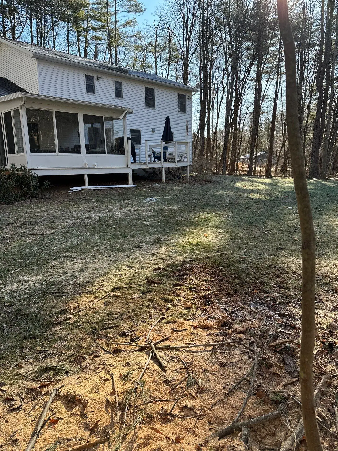A white two-story house with a screened porch and small deck overlooks a yard with patchy grass, bare soil, and scattered branches—ideal for improvement by experienced excavating contractors in Saratoga County, NY. Tall trees border the sunny backyard.