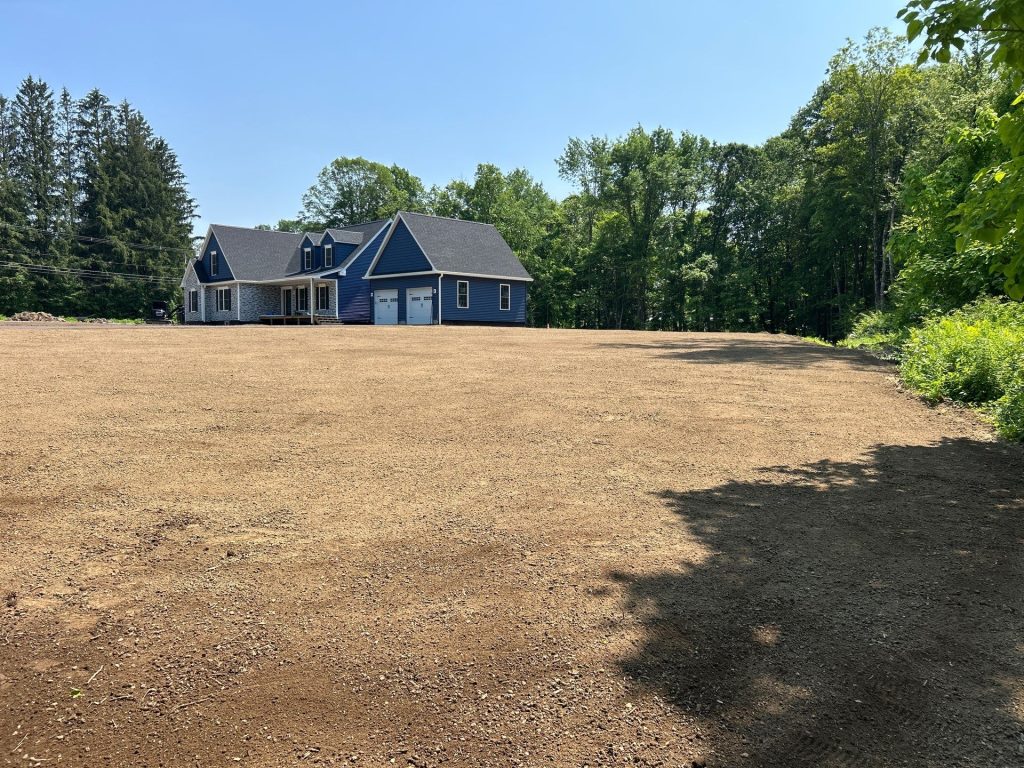 A newly constructed blue house with a front porch and white trim stands next to a large, cleared dirt lot—evidence of work by excavating contractors in Saratoga County, NY—surrounded by green trees under a clear blue sky.