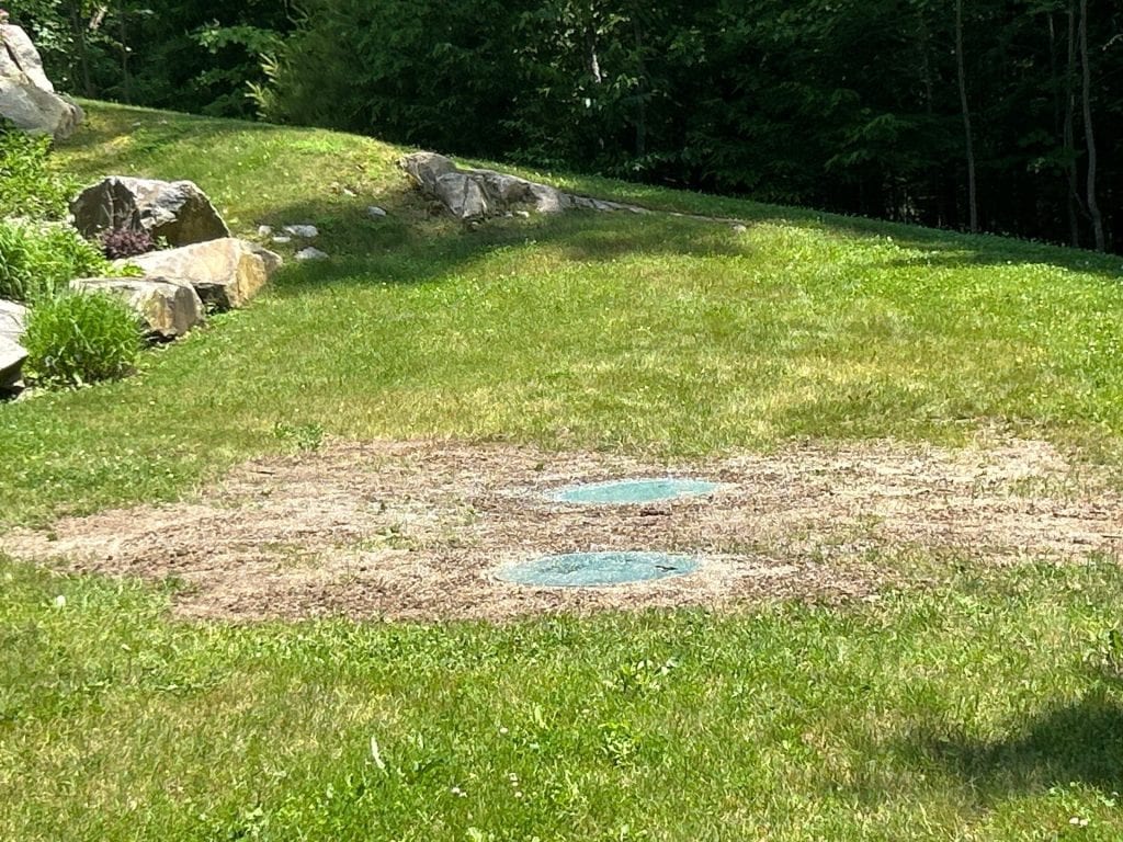 A grassy yard in NY with a large patch of bare soil in the center reveals two round, green septic tank covers—possibly recent work by excavating contractors Saratoga County. Large rocks and trees line the edge of the lawn in the background.