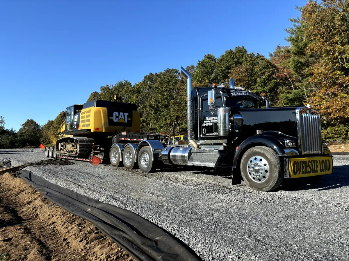 A black semi-truck with an "OVERSIZE LOAD" sign tows a large yellow CAT excavator for excavating contractors Saratoga County, NY, on a flatbed trailer along a gravel road, with trees and a clear blue sky in the background.