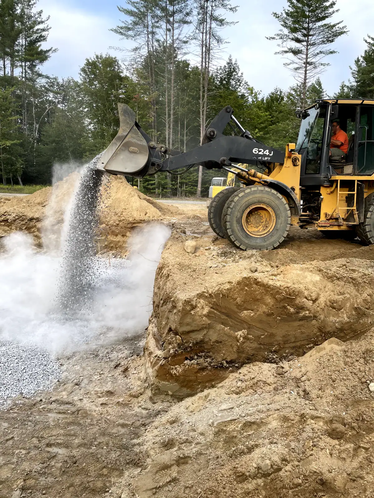 A yellow front loader operated by excavating contractors Saratoga County dumps gravel into a large pit at a NY construction site, creating a cloud of dust. Trees and mounds of earth are visible in the background.
