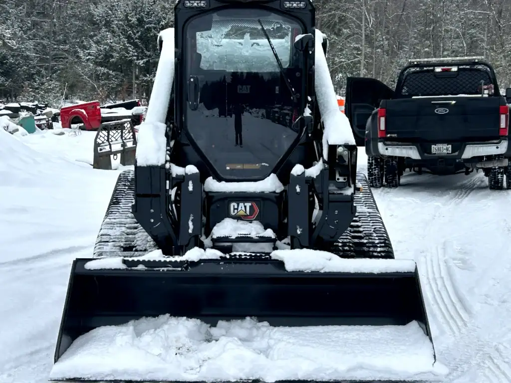 A black CAT skid steer loader, used by excavating contractors Saratoga County, NY, is covered in snow and parked on a snowy lot. Nearby, a black pickup truck with its driver door open sits as snow and trees fill the background.