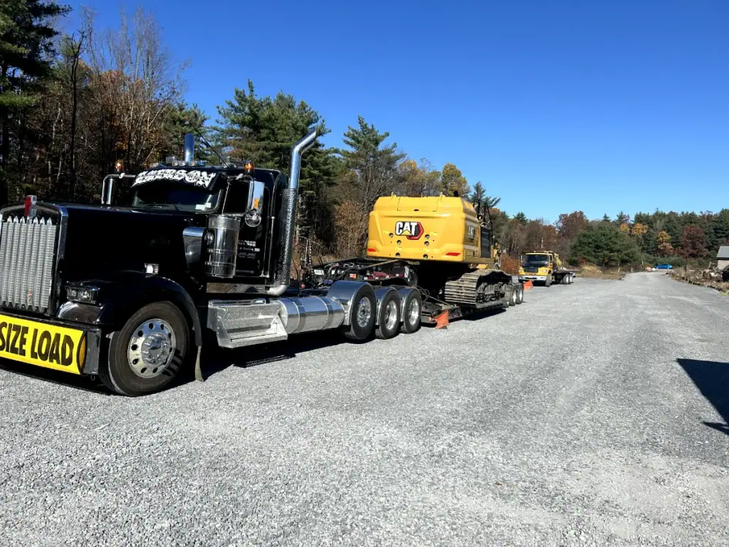 A large black semi-truck with an "OVERSIZE LOAD" sign hauls a yellow CAT excavator for excavating contractors Saratoga County, NY, along a gravel road, with trees and another truck in the background under a clear blue sky.
