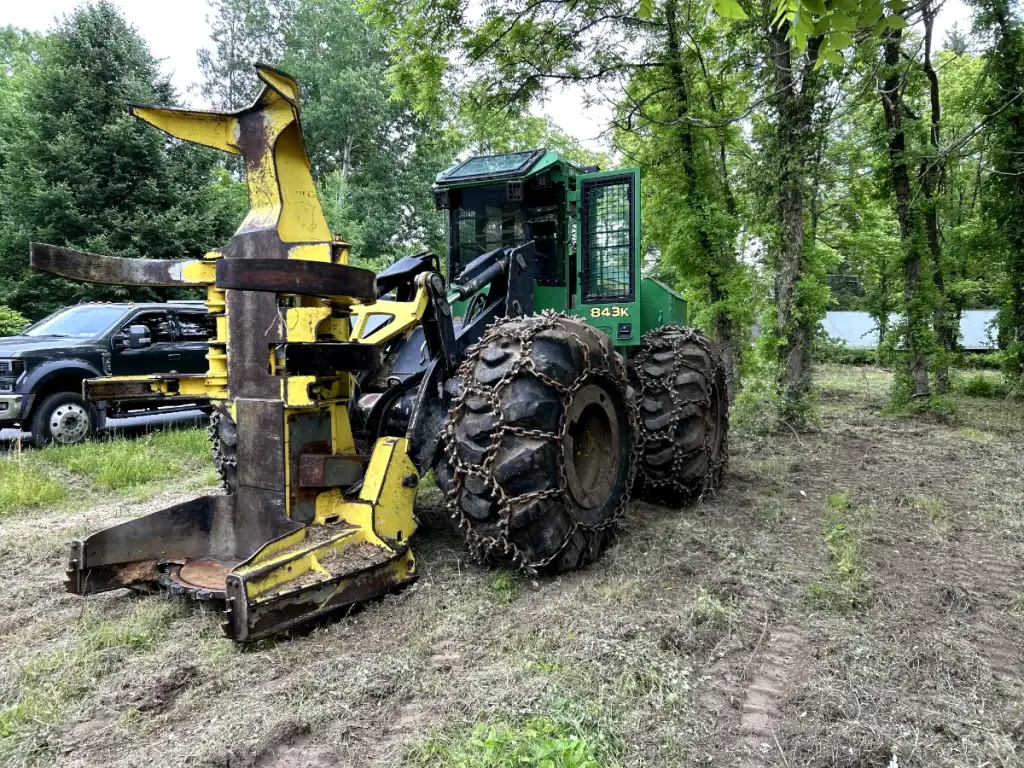 A large green forestry machine used by excavating contractors in Saratoga County, NY, with heavy chains on its tires and a yellow tree-grabbing attachment, is parked on cleared ground near trees, with a black pickup truck in the background.