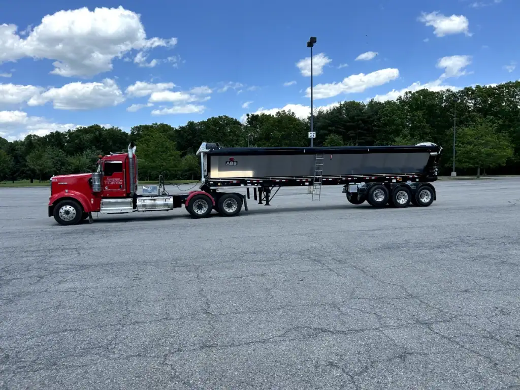 A red semi-truck with a large black dump trailer, used by excavating contractors Saratoga County NY, is parked on an empty asphalt lot, with green trees and a blue sky with scattered clouds in the background.
