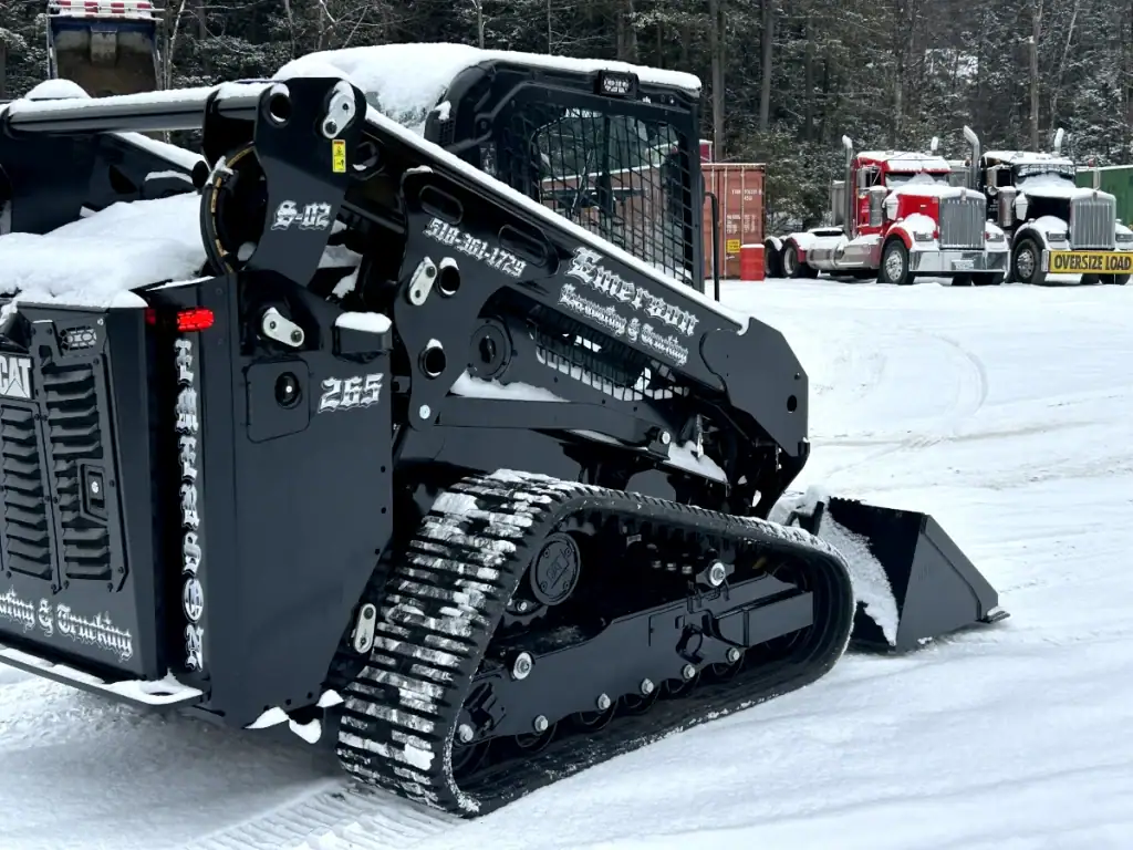 A black tracked skid steer loader with a snow-covered bucket sits on a snowy lot, surrounded by snow, containers, and a red semi-truck in the background—ideal equipment for excavating contractors in Saratoga County, NY.