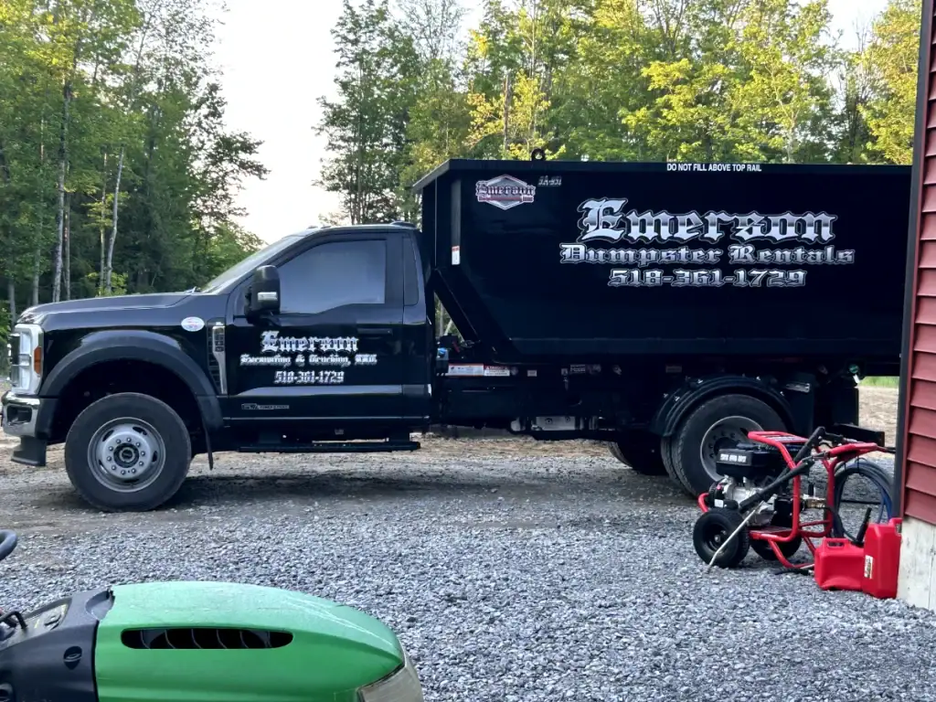 A black Emerson Dumpster Rentals truck, serving as one of the top excavating contractors Saratoga County, NY, is parked on a gravel driveway near trees. A red tricycle and green equipment are in the foreground, with contact info displayed in white lettering.