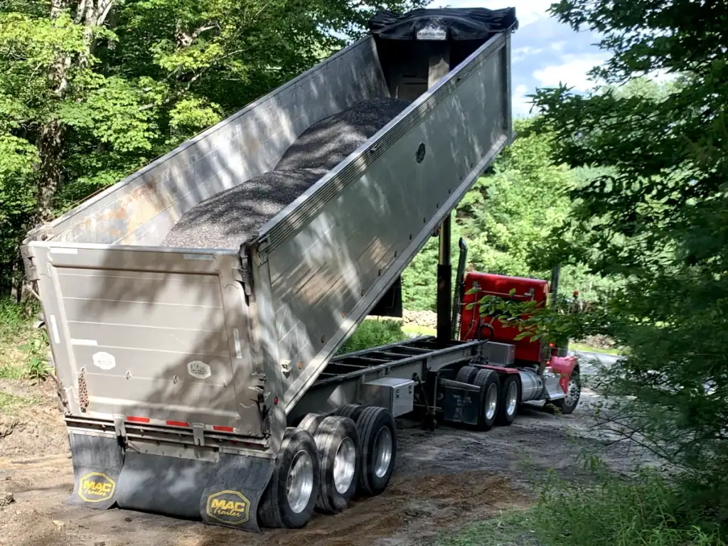 A red semi-truck with a gray dump trailer, often used by excavating contractors Saratoga County, NY, is unloading gravel onto a dirt road in a wooded area, with the trailer raised at a steep angle.
