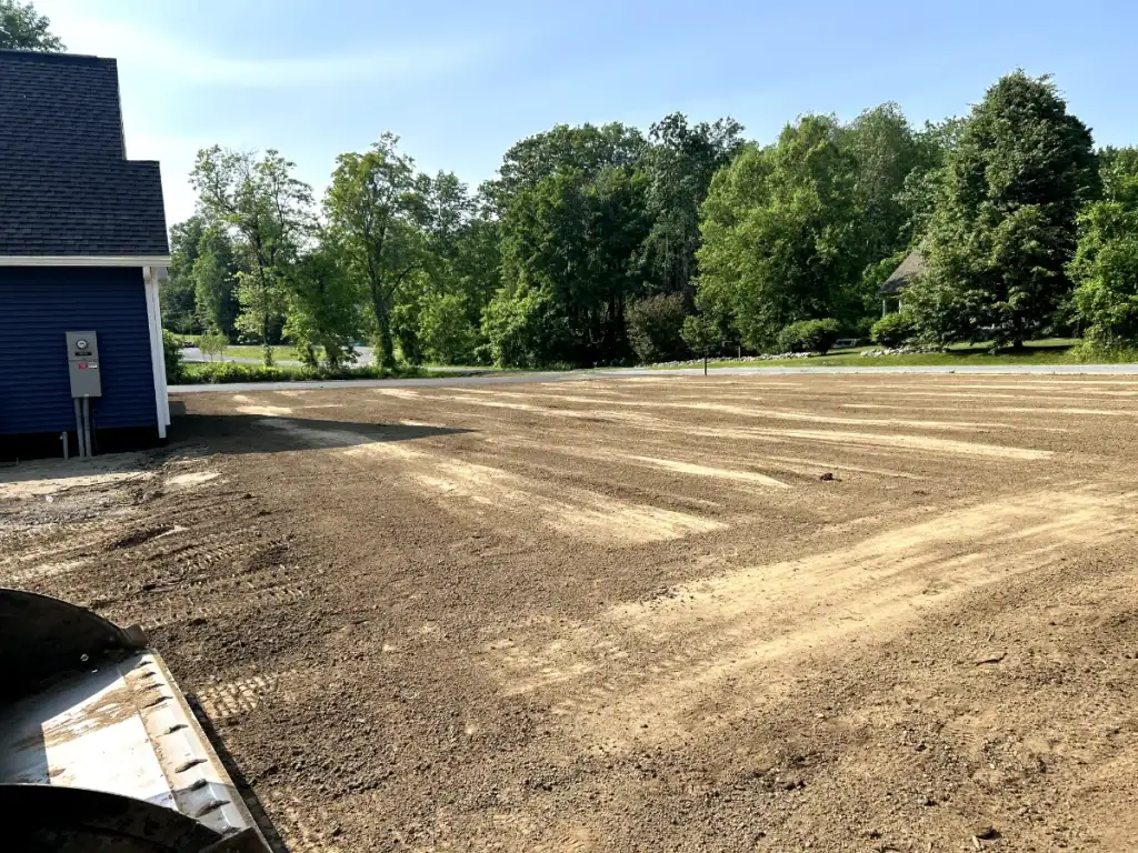 A freshly graded yard with bare soil next to a blue house in NY, bordered by green trees and a clear sky, ready for landscaping or seeding—expertly prepared by excavating contractors Saratoga County trusts.