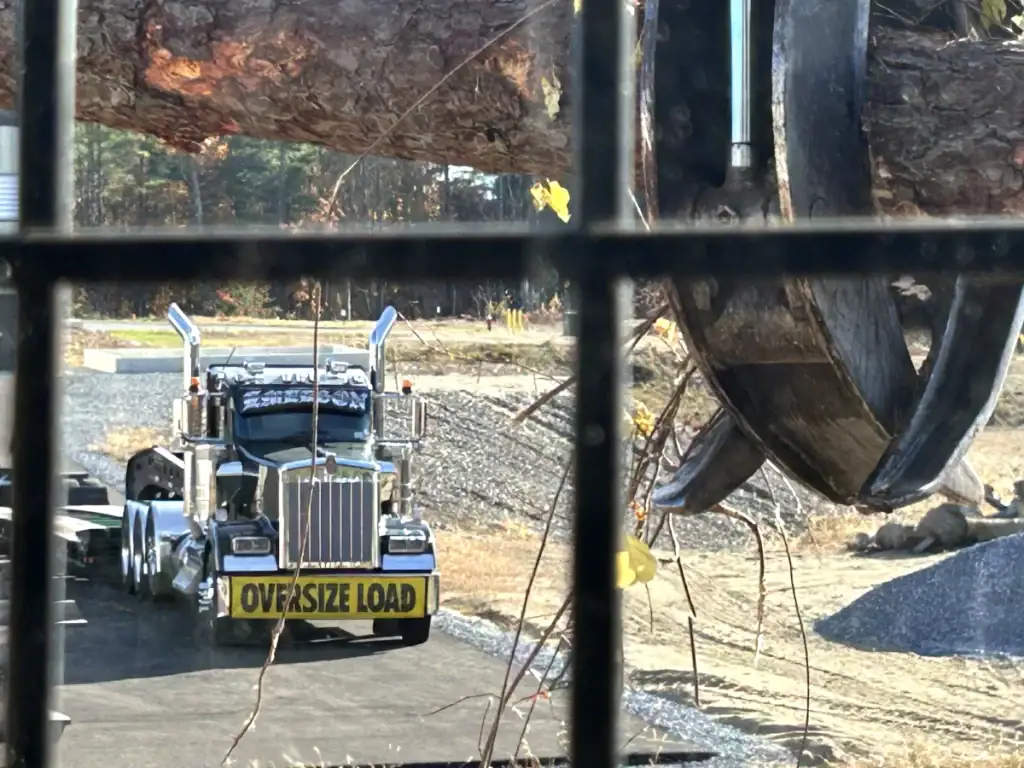 A large truck with an "OVERSIZE LOAD" sign is parked on a road, while a claw machine—often used by excavating contractors Saratoga County, NY—lifts a massive log nearby. The view is partially obscured by metal bars in the foreground.