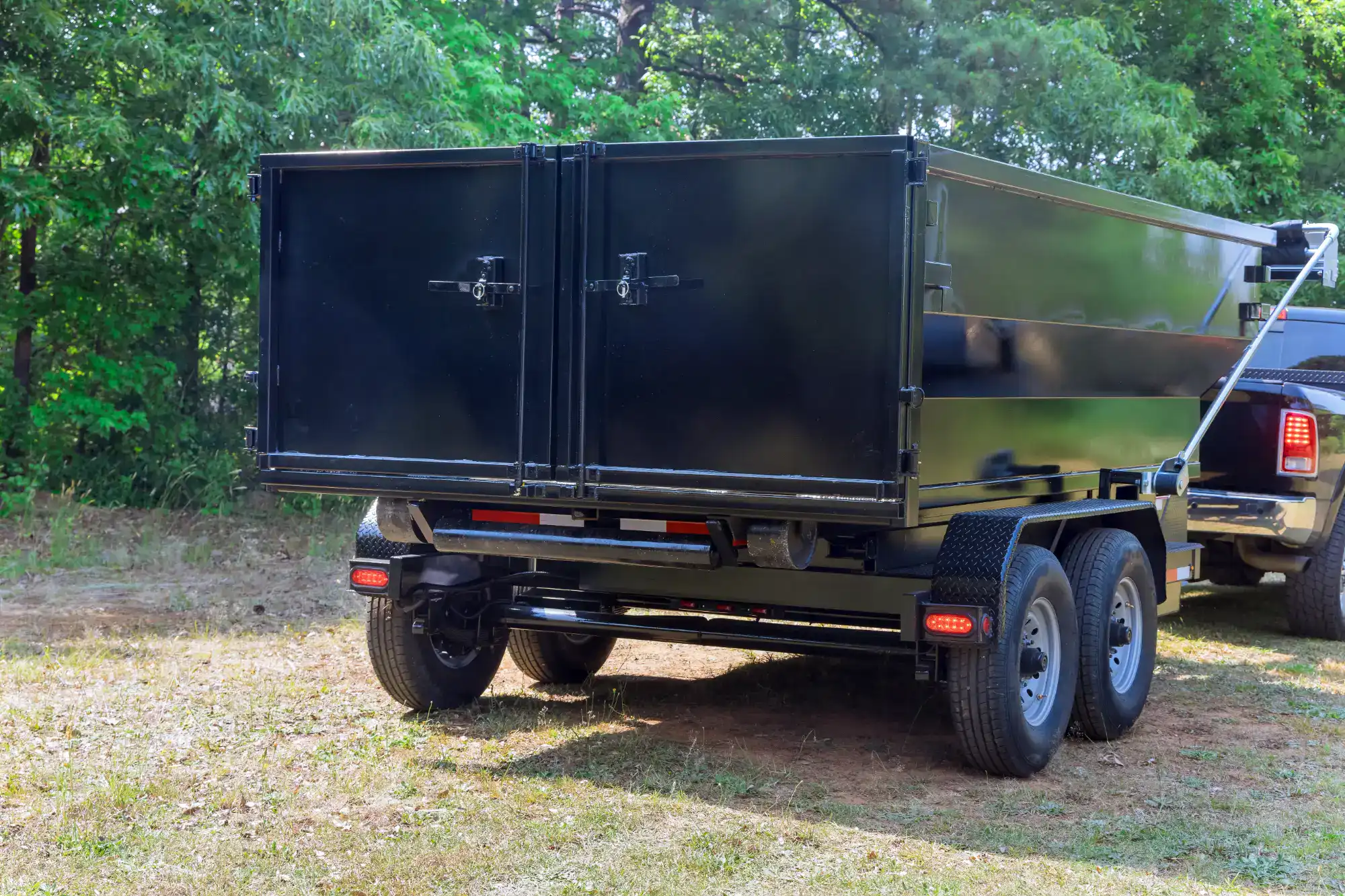 A black metal dump trailer with double rear doors is attached to a pickup truck and parked on grass, surrounded by green trees and foliage in the background.