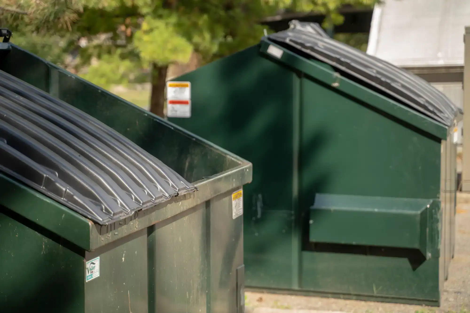 Two large green dumpsters with closed black lids are positioned outdoors on a sunny day, with trees and greenery visible in the background.