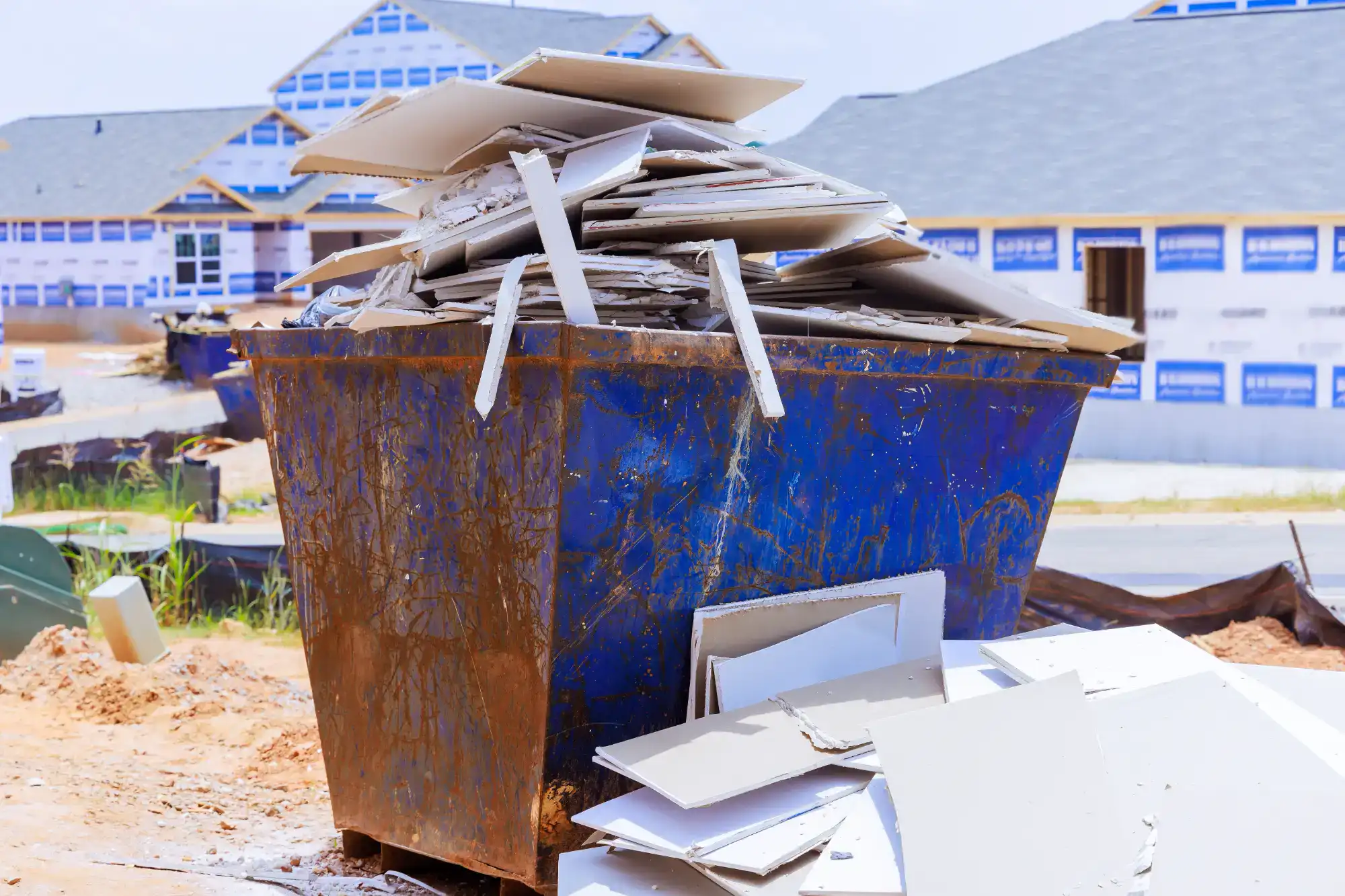 A large blue dumpster overflowing with broken drywall pieces sits at a construction site with partially built houses in the background.