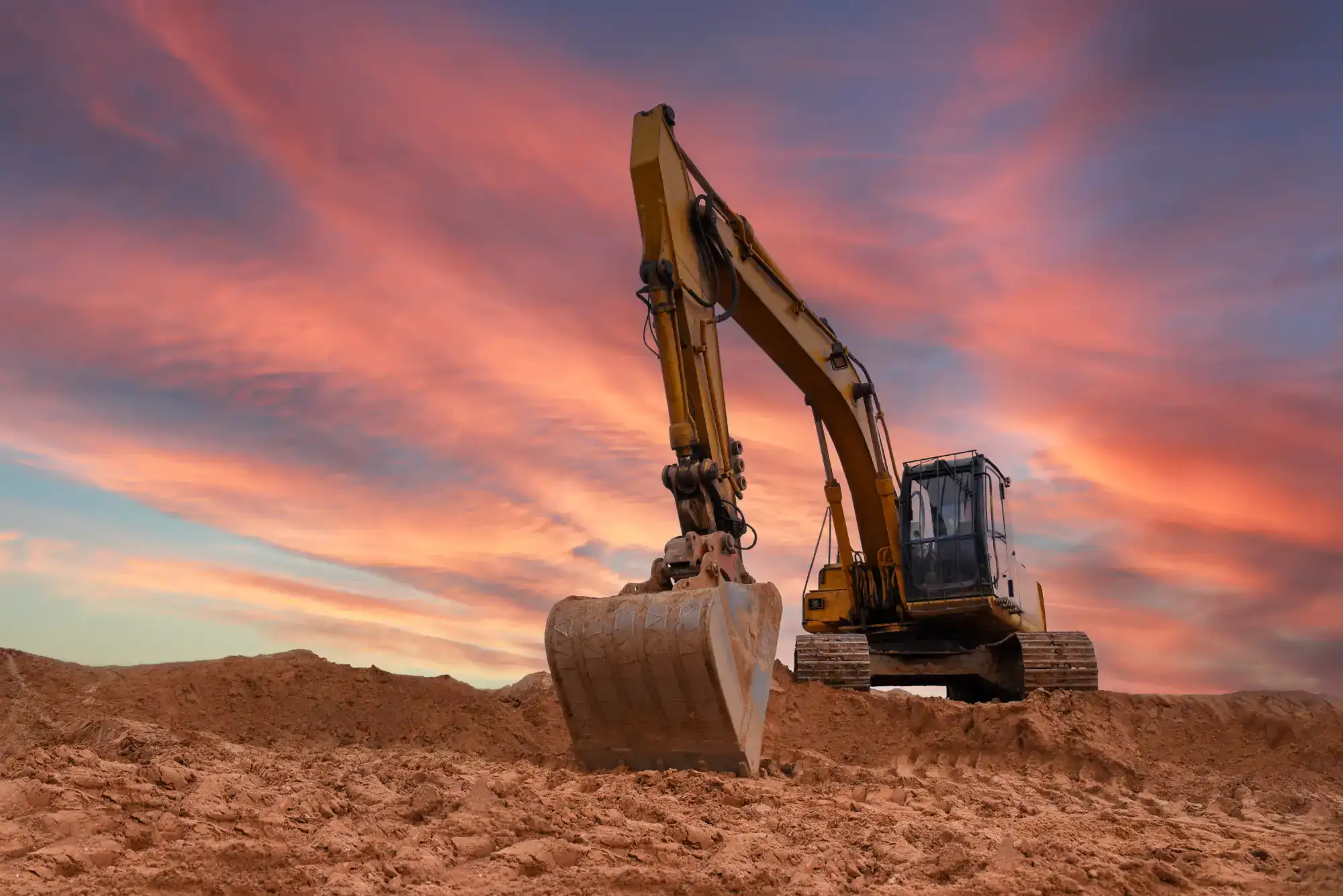 A large yellow excavator sits on a mound of dirt at a construction site, with a dramatic sunset sky of pink, orange, and blue hues in the background.