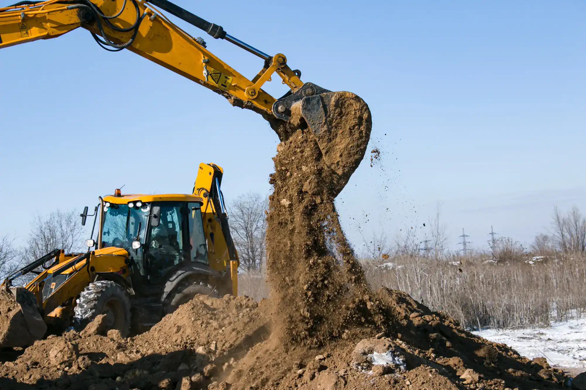 A yellow excavator lifts and pours a large amount of dirt onto a pile at a construction site, with bare trees and a clear blue sky in the background.
