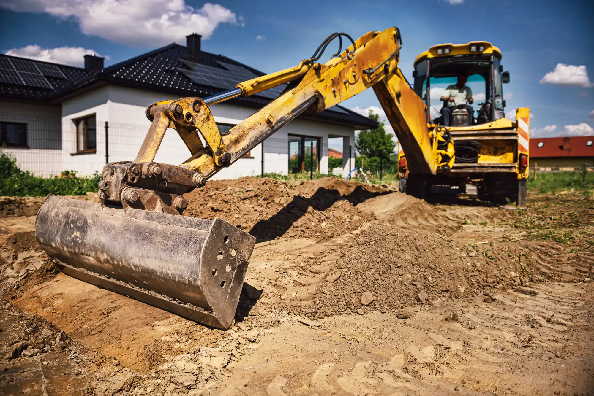 A yellow backhoe loader with its bucket raised is digging soil in front of a modern house under a sunny, blue sky with some clouds.