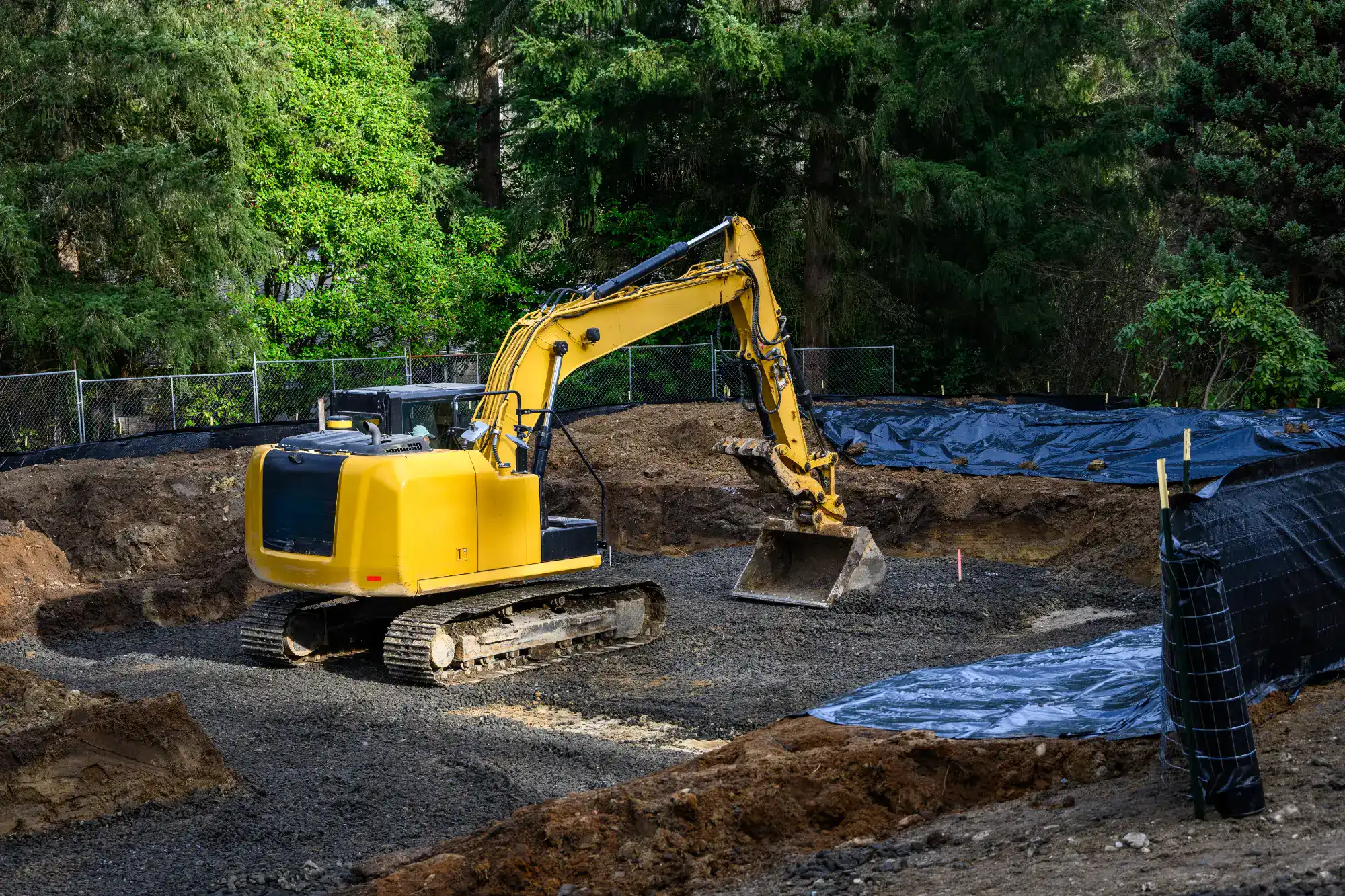 A yellow excavator sits on a gravel surface at a construction site surrounded by trees and fencing, with soil and a black tarp covering nearby areas.