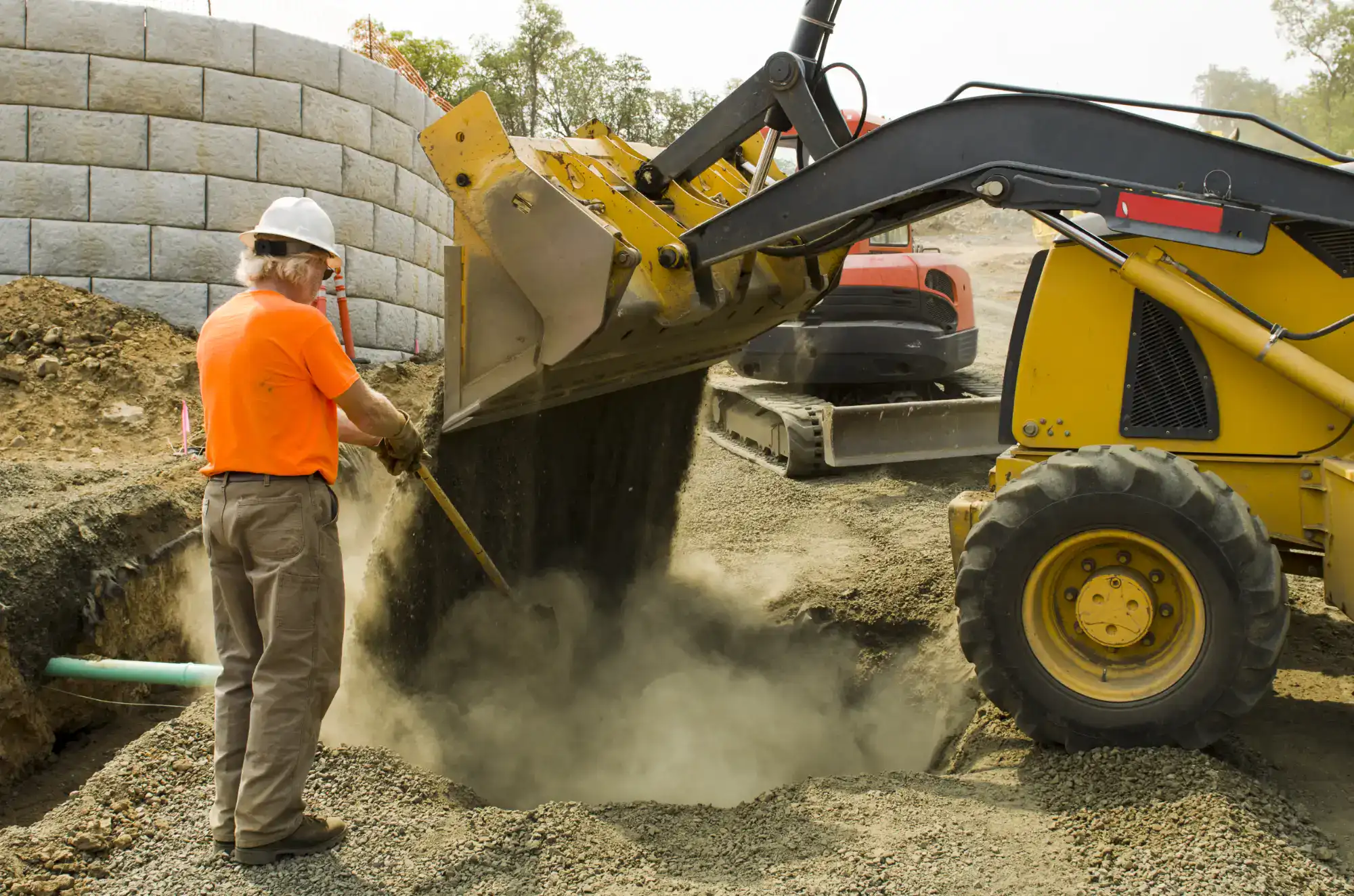 A construction worker in an orange shirt and white hard hat guides a yellow bulldozer as it dumps dirt into a trench at a construction site, with dust rising from the ground.