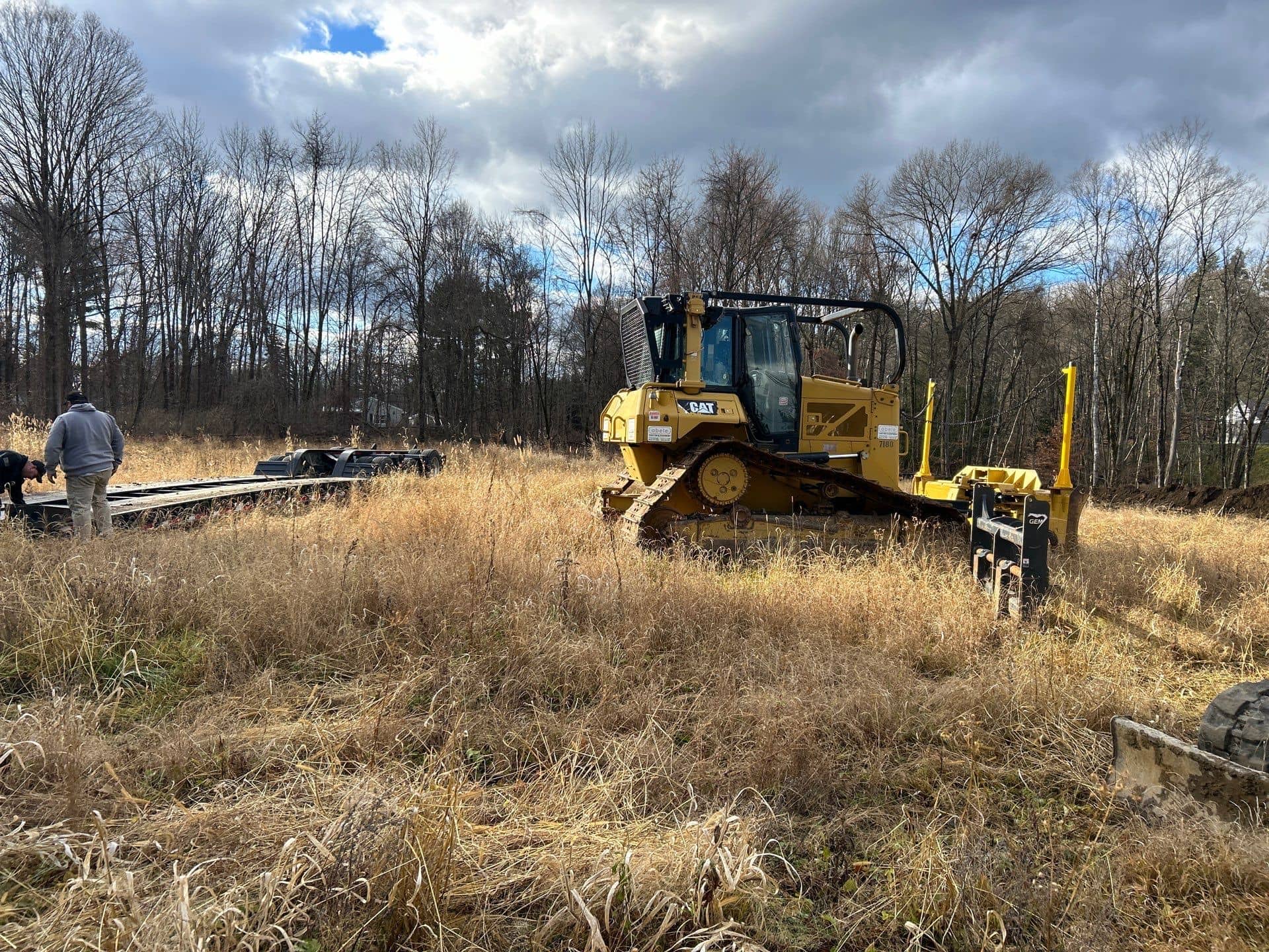 A yellow bulldozer from excavating contractors Saratoga County, NY sits in a grassy field with bare trees behind. A person stands near equipment on the left side under a cloudy sky.
