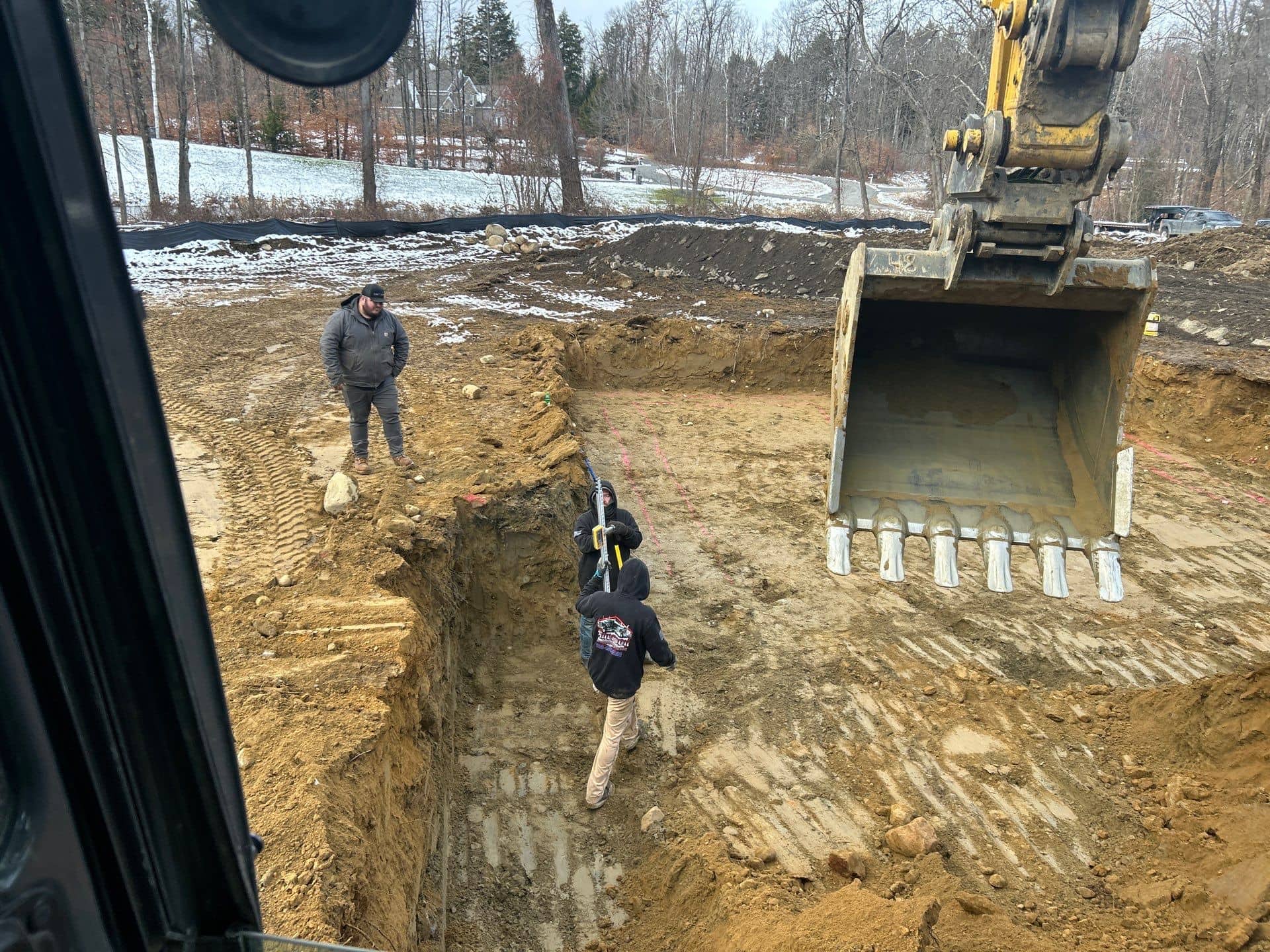 Two construction workers stand in a partially dug pit while a large yellow excavator bucket hovers nearby. Another person stands on the edge of the NY site, with bare trees and light snow in the background—excavating contractors Saratoga County at work.