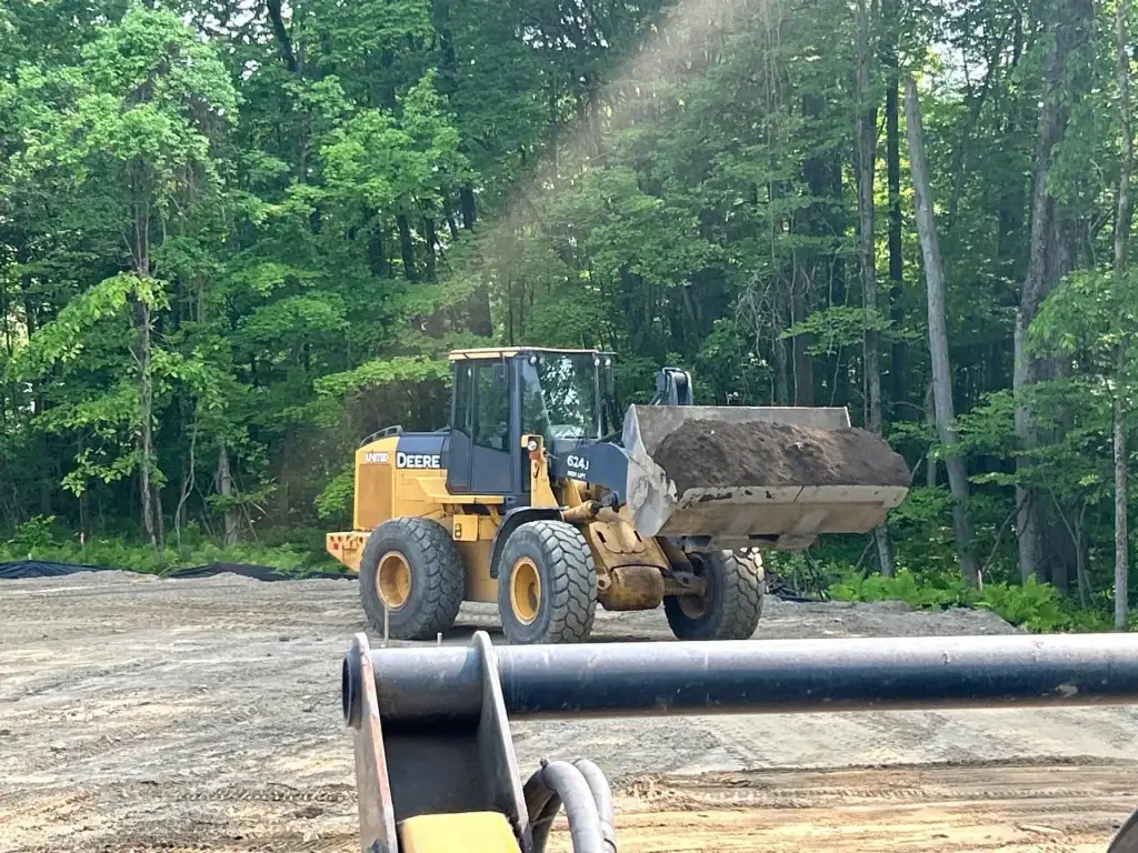 A yellow front-end loader with a filled bucket is on a cleared dirt area near a forest in NY, with trees and greenery in the background. The foreground shows part of another vehicle from excavating contractors Saratoga County.