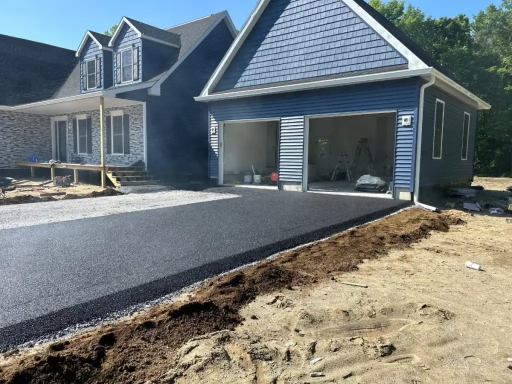 A newly paved asphalt driveway leads up to a blue house with an open garage. Construction materials from NY excavating contractors Saratoga County are visible inside, and the surrounding yard remains unfinished with exposed dirt.
