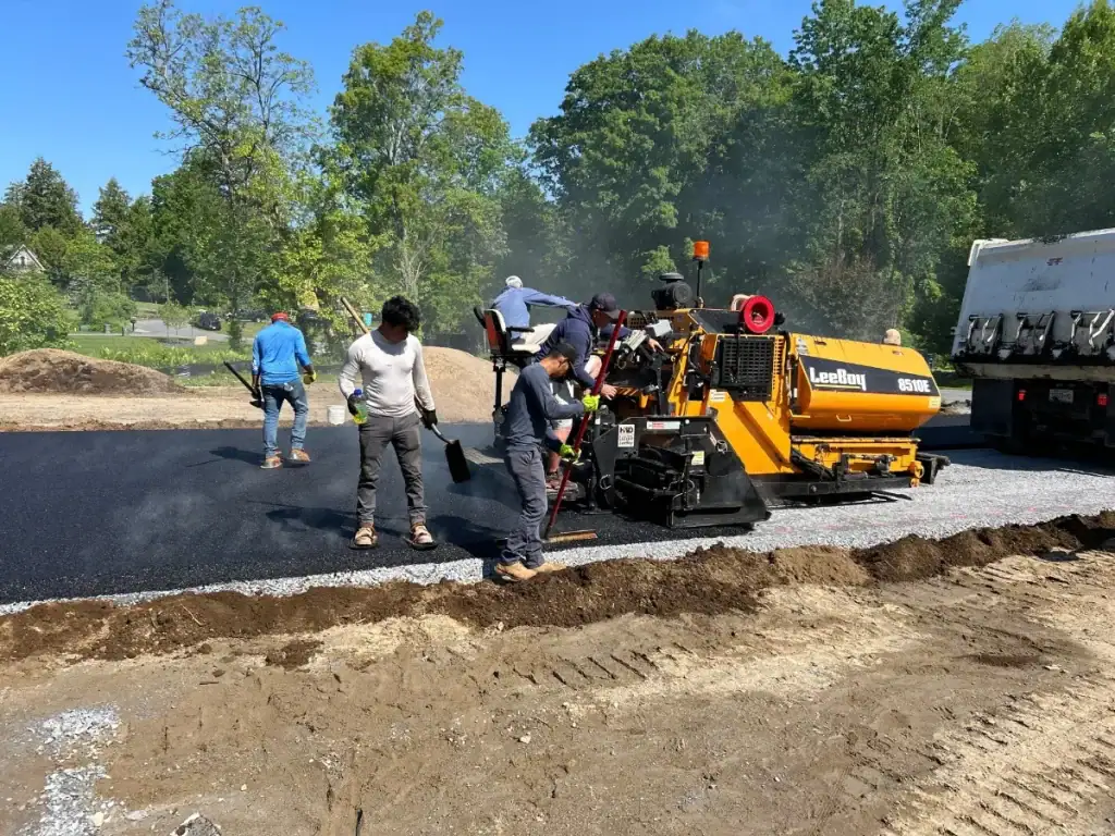 Workers operate machinery and use shovels to spread and smooth fresh asphalt on a road construction site, while experienced excavating contractors Saratoga County, NY ensure quality results under blue skies on a sunny day.