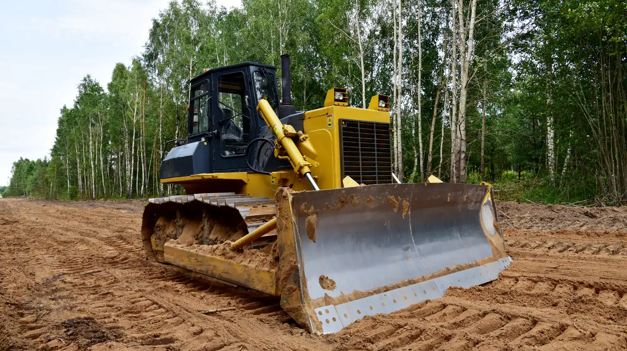 A yellow bulldozer with a large metal blade is parked on a dirt path in a forest clearing, surrounded by green trees and a clear sky in the background.