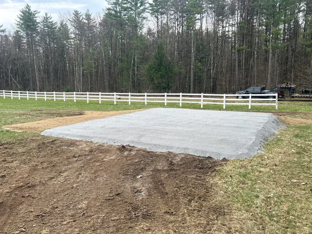 A rectangular gravel pad sits on a grassy field near a white fence in NY, with a forest of tall trees and a black pickup truck in the background—perfect work by experienced excavating contractors Saratoga County trusts. The ground is slightly uneven and muddy.