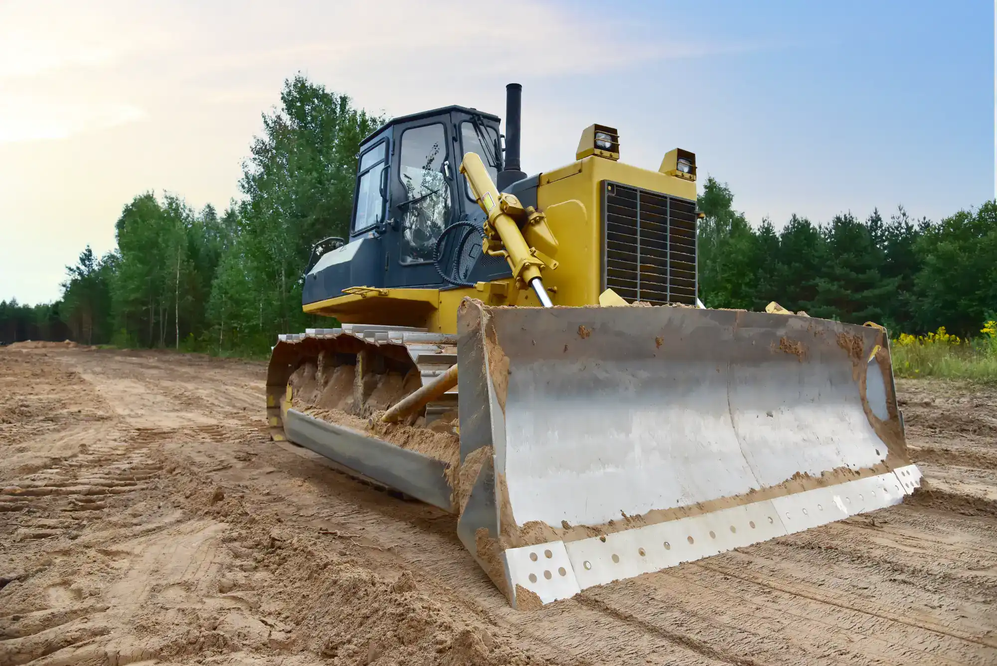 A yellow bulldozer with a large front blade stands on a dirt path, surrounded by trees under a clear sky, ready for construction or earth-moving work.