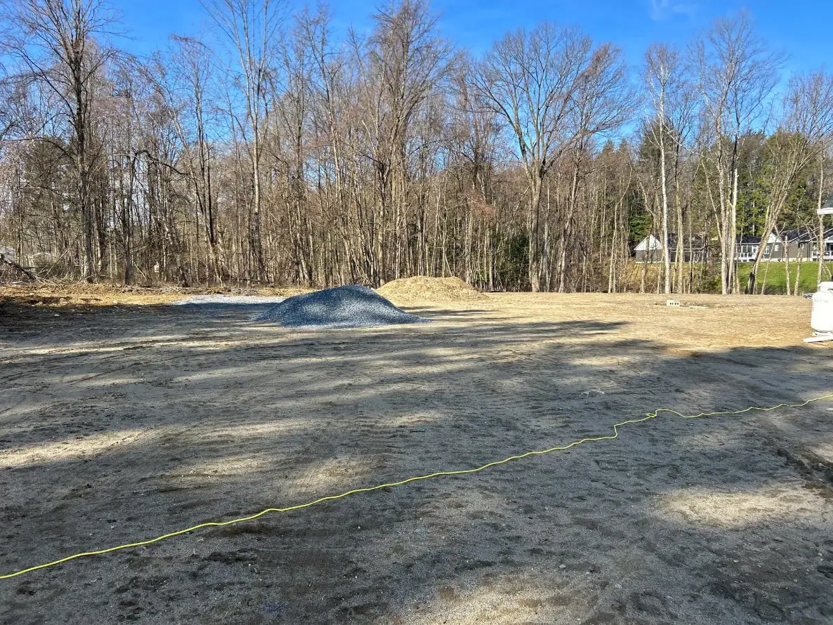 A cleared, dirt construction site in NY bordered by leafless trees, with piles of gravel and sand in the center—excavating contractors Saratoga County at work—while a yellow extension cord stretches along the ground under a bright blue sky.