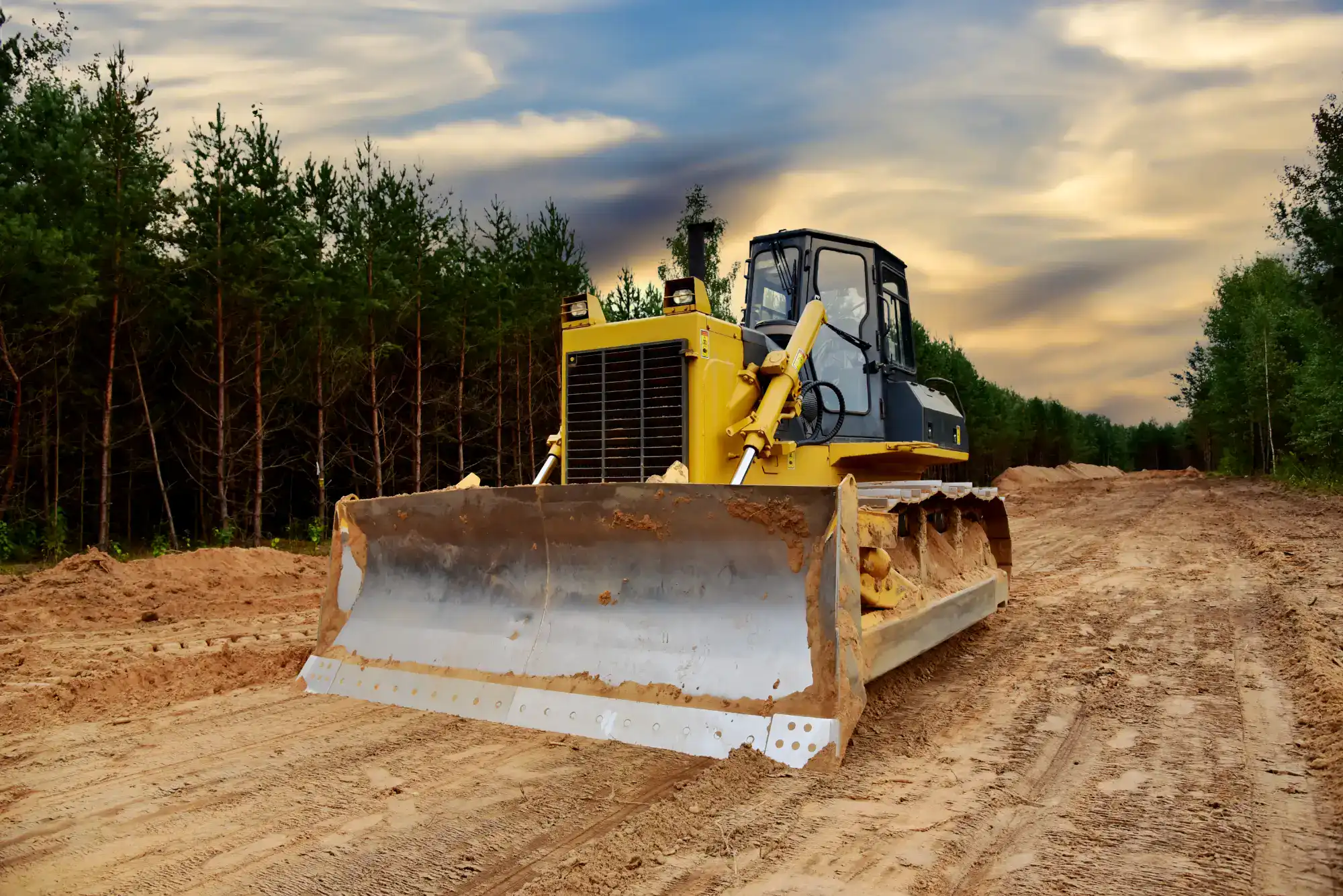A yellow bulldozer on a sandy dirt road surrounded by trees, with a dramatic cloudy sky at sunset in the background.