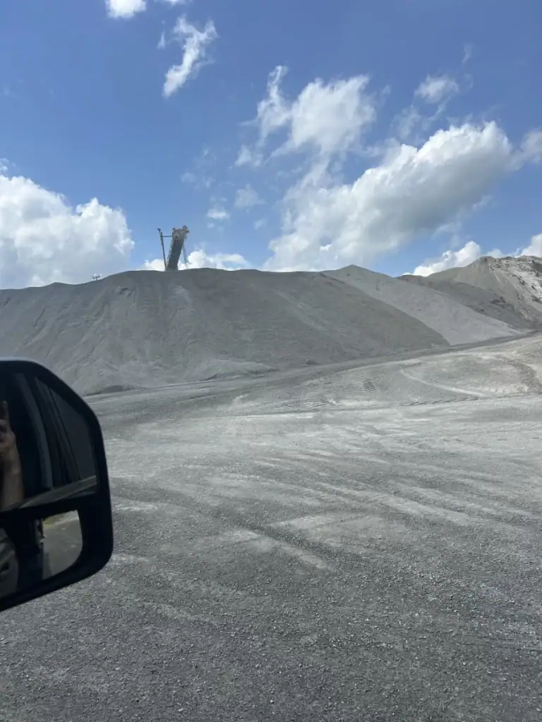 A large gravel mound sits under a partly cloudy blue sky, with industrial equipment visible on top. The photo is taken from inside a vehicle, with a side mirror visible in the lower left corner.