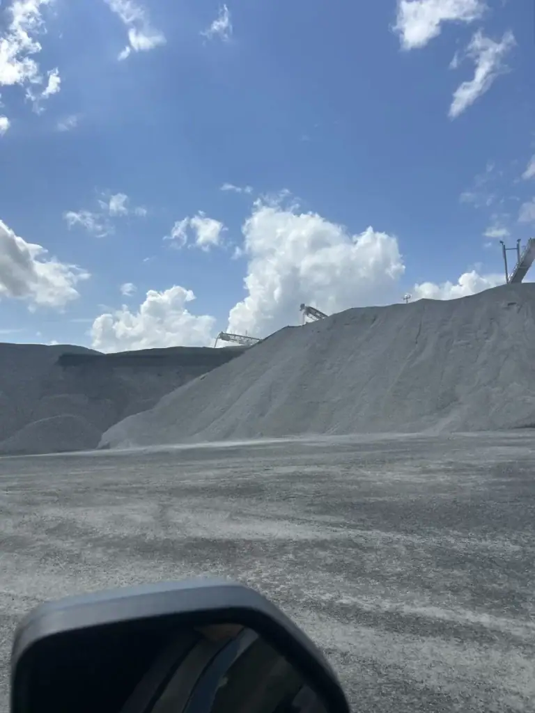 Large mounds of gray gravel or sand under a bright blue sky with scattered clouds; industrial equipment and conveyor belts are visible on top of the piles, and part of a car mirror is seen in the foreground.