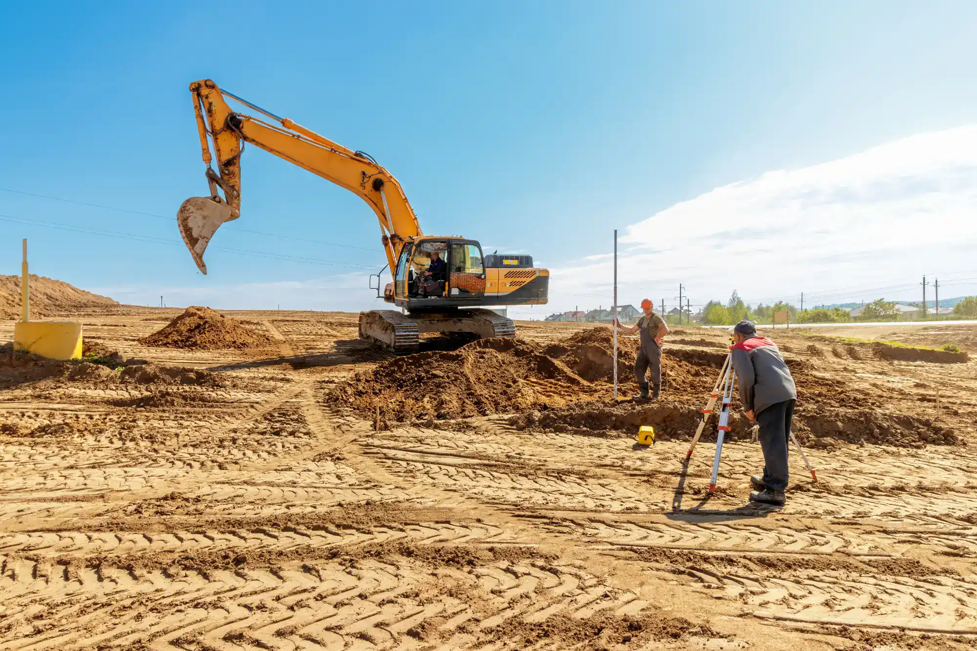 Two construction workers survey a sandy construction site with a yellow excavator digging in the background under a clear blue sky. Tire tracks and piles of dirt are visible on the ground.