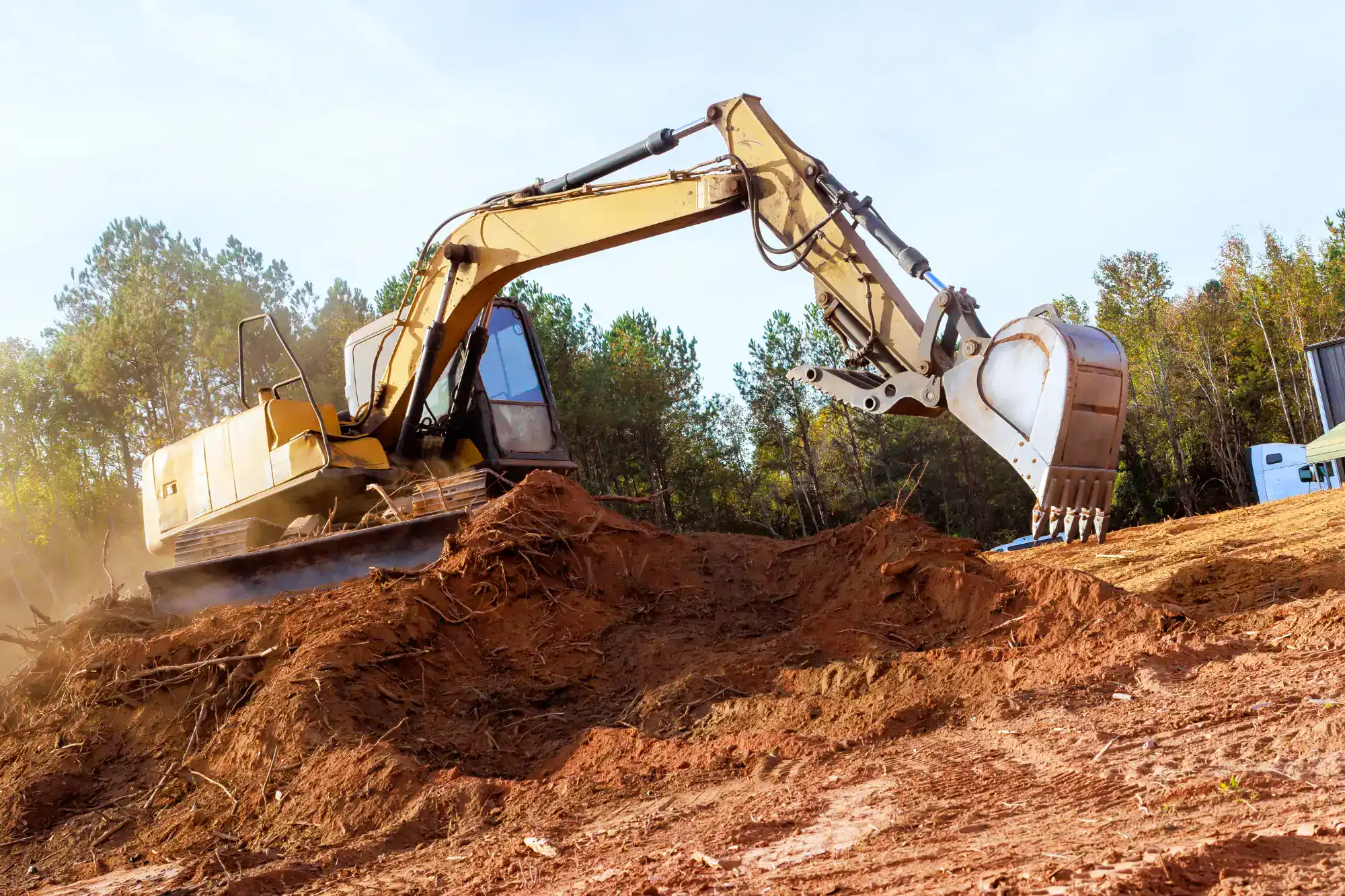 A yellow excavator is digging and moving earth on a construction site, with soil and dirt piled around. Trees and clear sky are visible in the background.