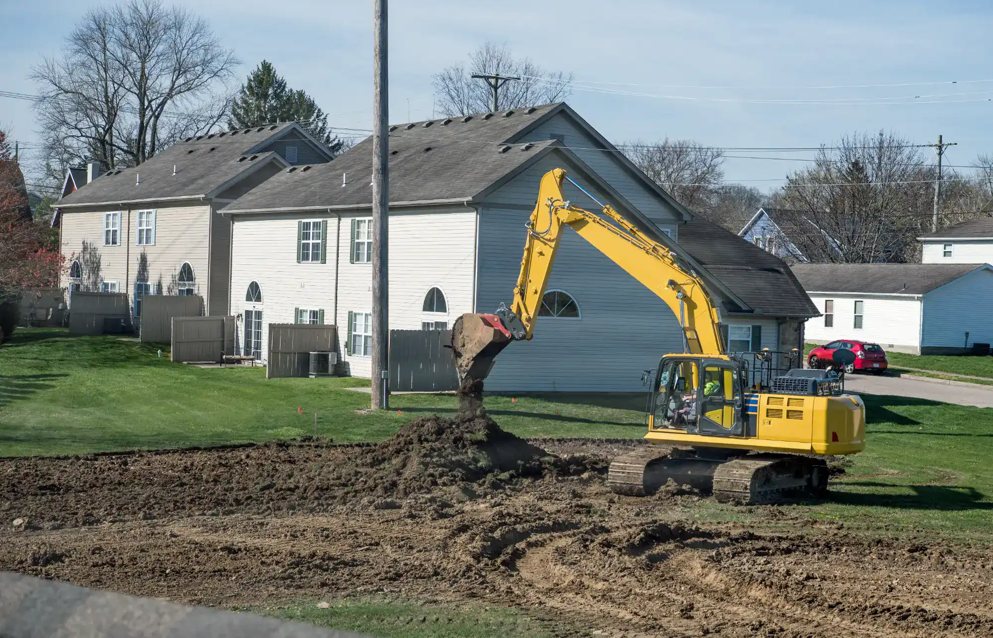 A yellow excavator lifts dirt on a grassy lot near a row of light-colored houses, preparing land for construction on a clear day.