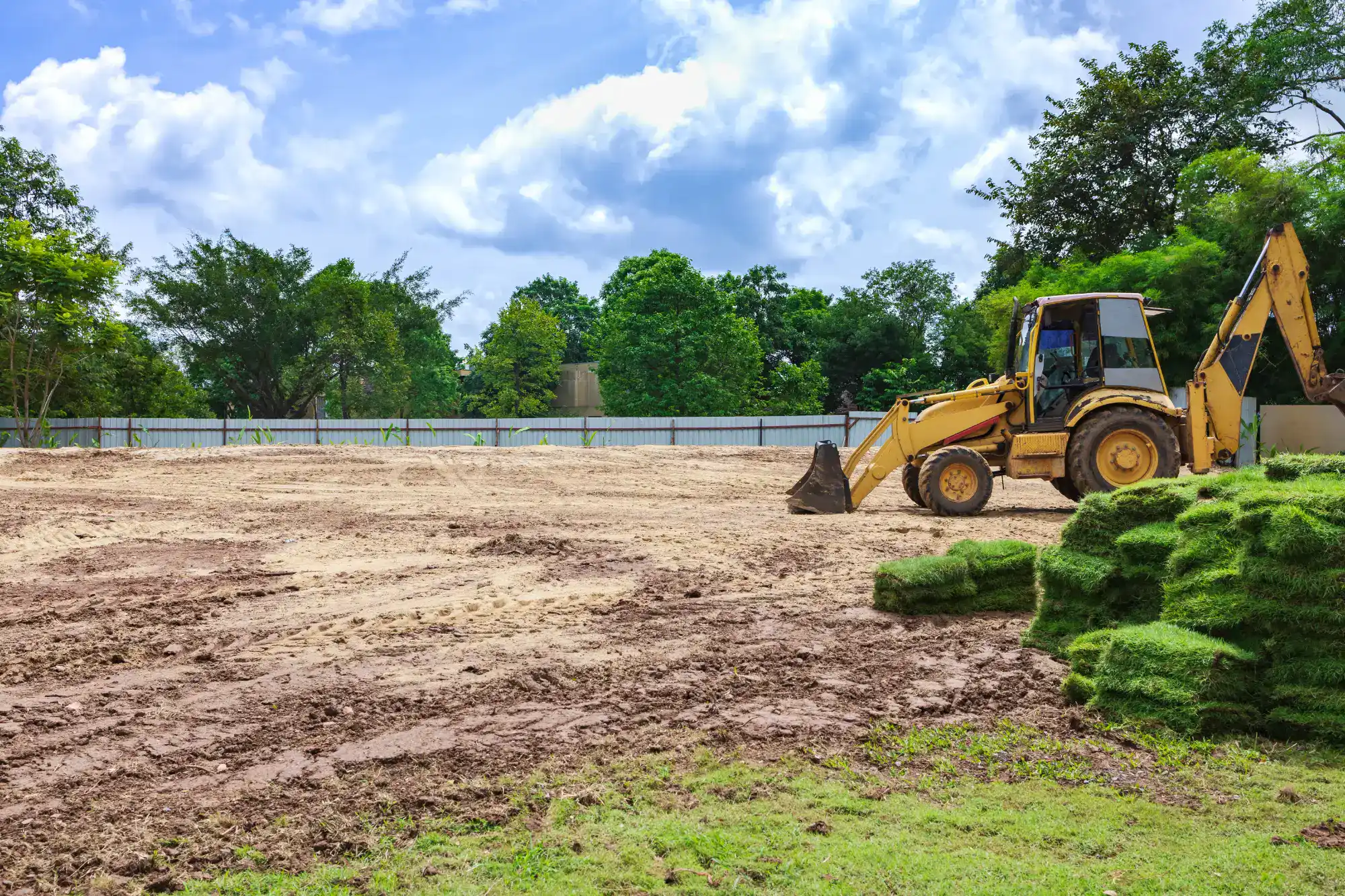 A yellow backhoe loader is parked on a cleared, dirt-covered construction site with stacks of rolled sod in the foreground and trees in the background under a partly cloudy sky.