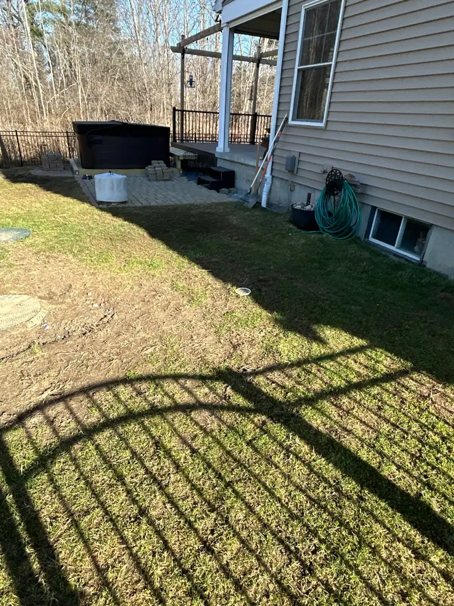 A backyard in NY with patchy grass, a garden hose coiled on the ground, a black hot tub on a paver patio, some containers, and a fence casting shadows across the lawn—perfect for ideas from excavating contractors Saratoga County trusts.